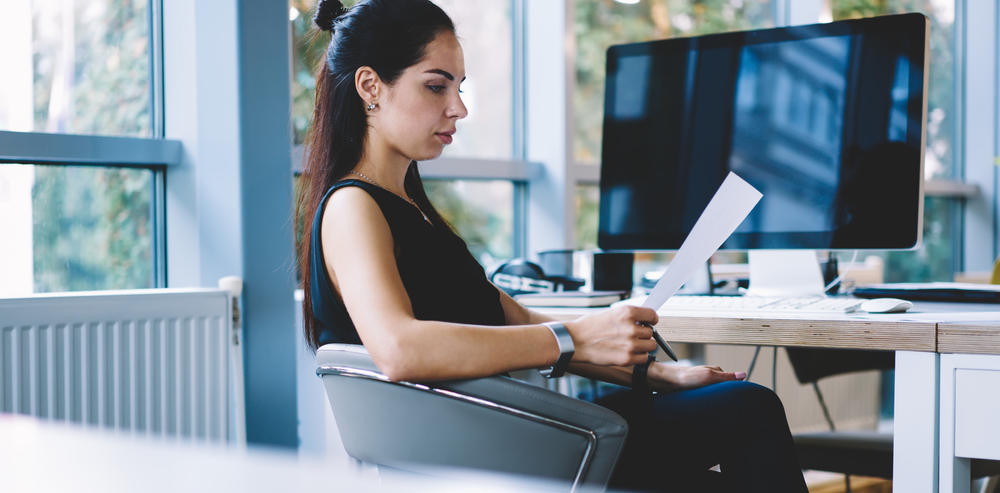 Side view of concentrated businesswoman in stylish formal clothes sitting at office table with laptop and reading documents in daytime